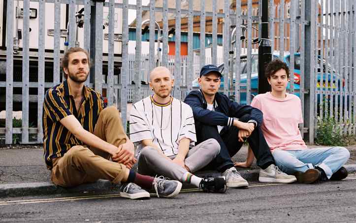 Four male band members sat on the curb in front of a tall fence.