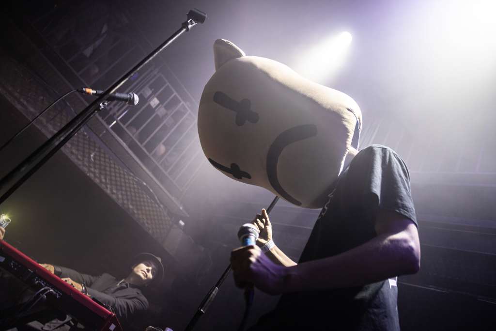 Shadowed figure holds a microphone on stage while a keyboard player sits behind in a trilby hat.