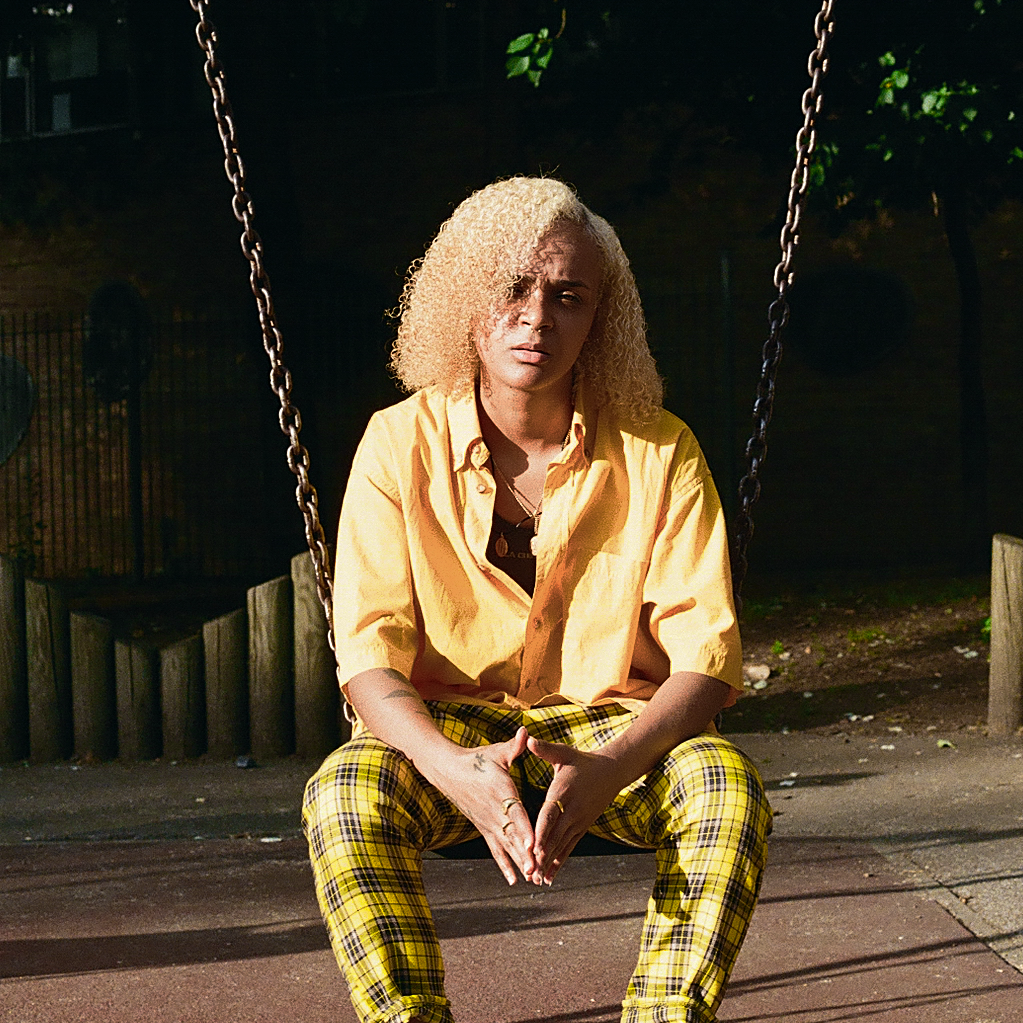 Nicki Knightz in a yellow shirt and checkered yellow trousers, sat on a swing.