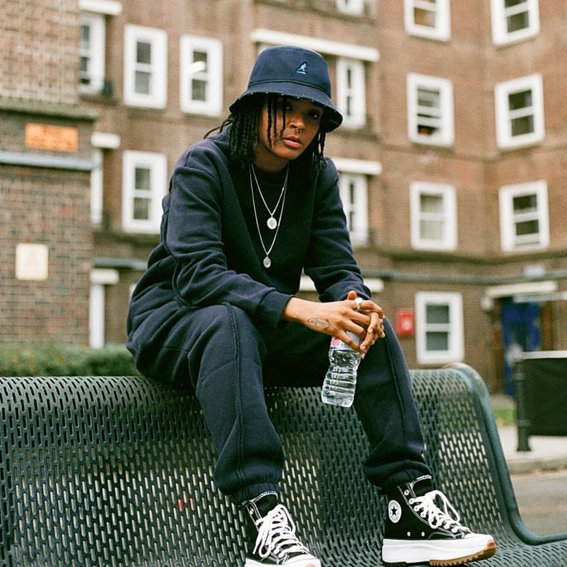 Nicki Knightz sat on a bench in a Kangol bucket hat outside a housing estate.